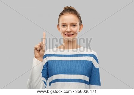 gesture, count and people concept - smiling teenage girl in pullover pointing one finger up over grey background