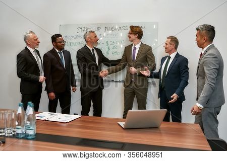 Two Happy Businessmen In Classic Wear Shaking Hands While Having A Meeting With Their Team In The Mo