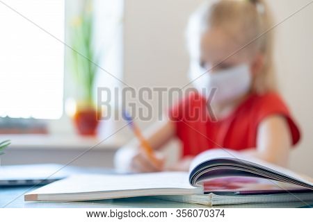 Blurred Blonde Schoolgirl In Medical Mask Studying At Home, Doing School Homework, Writing In Notebo