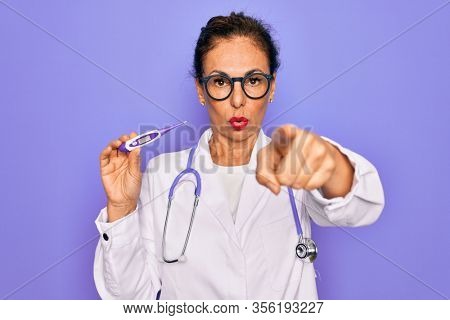 Middle age senior professional doctor woman holding medical thermometer pointing with finger to the camera and to you, hand sign, positive and confident gesture from the front
