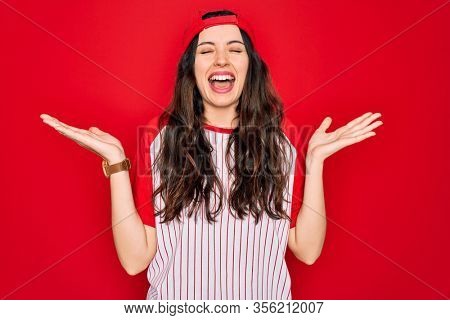 Beautiful woman with blue eyes wearing baseball sportswear and cap over red background celebrating mad and crazy for success with arms raised and closed eyes screaming excited. Winner concept