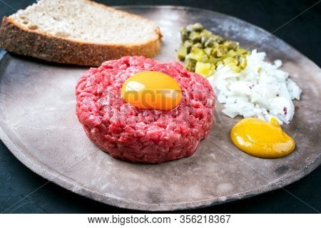Gourmet tartar raw from beef fillet with yellow of the egg, gherkin and farmhouse bread as closeup on a modern design rustic plate 