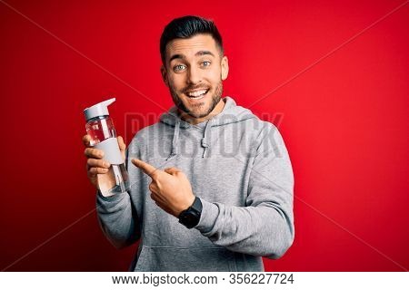 Young handsome sportsman drinking bottle of water to refeshment over red background very happy pointing with hand and finger
