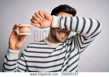 Young handsome man with beard holding plastic denture teeth over white background Smiling cheerful playing peek a boo with hands showing face. Surprised and exited