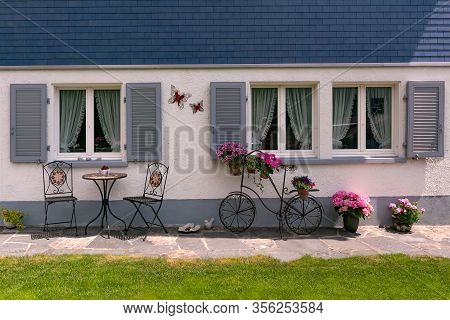 Facade Of Modern Swiss House With Sitting Corner And Flower Pots, Decorative Forged Bicycle In Swiss