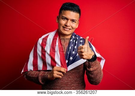 Young handsome latin patriotic man wearing united states of america falg happy with big smile doing ok sign, thumb up with fingers, excellent sign