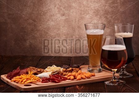 Three glasses with different beer and set of various kind snacks: jerky,  potato chips, salted nuts, stockfish, bread spicy crackers, dried squids on wooden table on brown background. Snacks for beer.