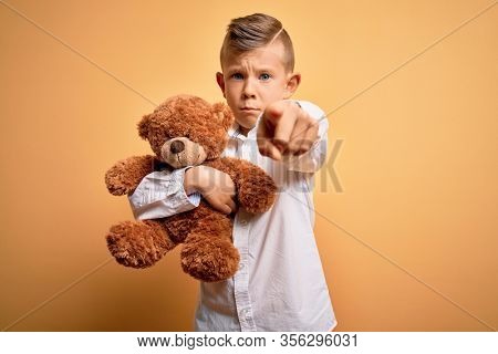 Young little caucasian kid hugging teddy bear stuffed animal over yellow background pointing with finger to the camera and to you, hand sign, positive and confident gesture from the front