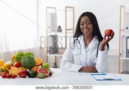 African American Smiling Female Doctor Dietologist Sitting At Workdesk, Holding Apple In Her Hand, E