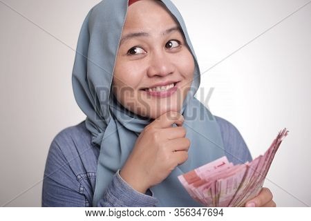 Portrait Of Indonesian Muslim Woman Holding Rupiah Money, Smiling And Thinking Gesture