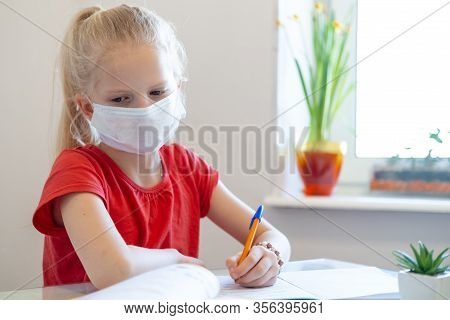Serios Sad Blonde Schoolgirl In Medical Mask Stydying At Home, Doing School Homework, Writing In Not