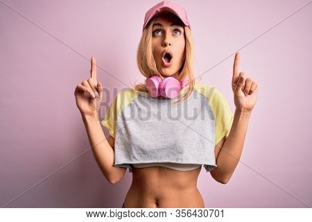 Young beautiful blonde sportswoman doing sport using headphones over pink background amazed and surprised looking up and pointing with fingers and raised arms.