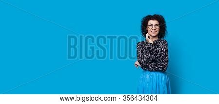 Cheerful Caucasian Lady With Curly Hair Smiling While Touching Her Chin On A Blue Background With Fr