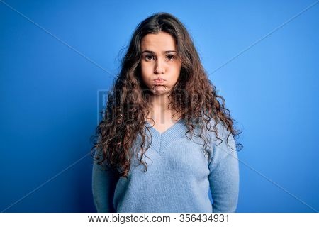 Young beautiful woman with curly hair wearing blue casual sweater over isolated background puffing cheeks with funny face. Mouth inflated with air, crazy expression.
