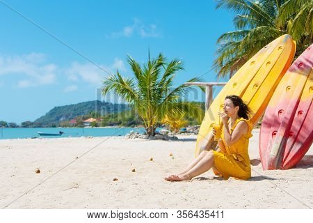 Girl In A Yellow Dress On A Tropical Sandy Beach Works On A Laptop Near Kayaks And Drinks Fresh Mang