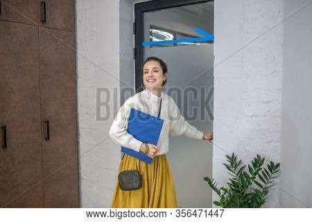 Young Woman Standing At The Door, Holding Door Hangle, Smiling