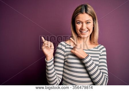 Young beautiful blonde woman wearing casual striped shirt over purple isolated background smiling and looking at the camera pointing with two hands and fingers to the side.