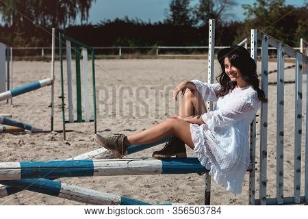 Beautiful Sexy Girl Posing On The Horizontal Crosspiece On A Horse Epodrom. Female Wearing White Dre