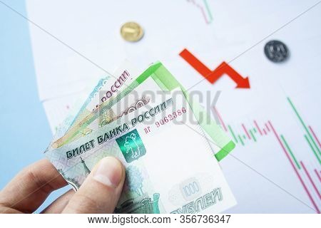 Male Hand Holding Rouble Bills On Exchange Rate Of Russian Ruble And Red Arrow. Ruble Depreciation. 