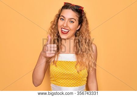 Beautiful blonde woman pin-up with blue eyes wearing red sunglasses over yellow background happy with big smile doing ok sign, thumb up with fingers, excellent sign