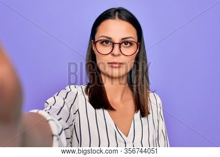 Young beautiful brunette woman wearing striped shirt and glasses making selfie by camera thinking attitude and sober expression looking self confident