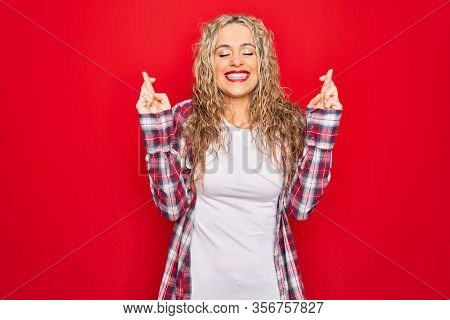 Young beautiful blonde woman wearing casual shirt standing over isolated red background gesturing finger crossed smiling with hope and eyes closed. Luck and superstitious concept.