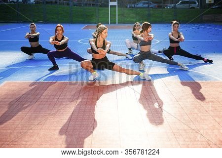 Group Of Attractive Young Athletic Girls In Sportswear Doing Exercises With Squats On The Sportgroun