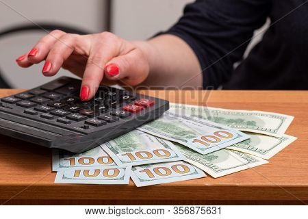 Womans Hands, Consider On The Calculator Hundred Dollar Bills Over The Table. Selective Focus