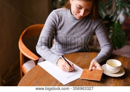 Young Cauacsian Girl Filling Visa Application At Cafe, Passport On Table.