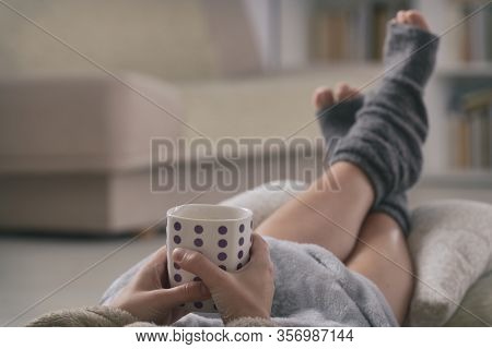 Woman with cup of hot drink  and feet lying on pilow at home. Quarantine and isolation concept