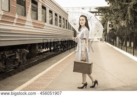 An Elegant Lady In A Dress With An Umbrella And A Vintage Suitcase Is Waiting For Her Train On The S