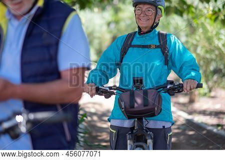 Active Senior Caucasian Couple With Electrobikes Standing Outdoors In The Park. Two Happy Elderly Pe