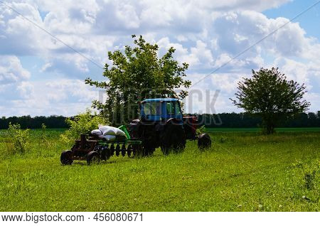 Trailed Old Harrow With Agricultural Tractor Against The Background Of Green Field On An Summer Day.