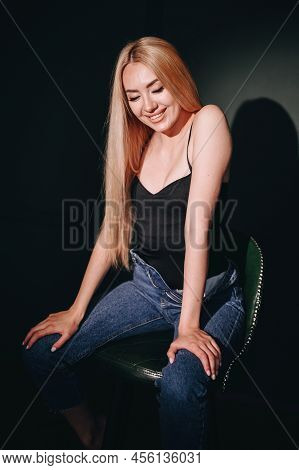 Girl With Long Blond Hair With Unbuttoned Jeans And A Black T-shirt Is Sitting On A Bar Stool, Smili