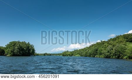 A Calm Blue River Under An Azure Sky. Lush Green Vegetation On Hilly Shores. White Clouds. Copy Spac