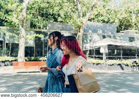 Young Asian Woman In Casual Clothes With Paper Bags Holding Arm Of Best Friend And Walking On City S