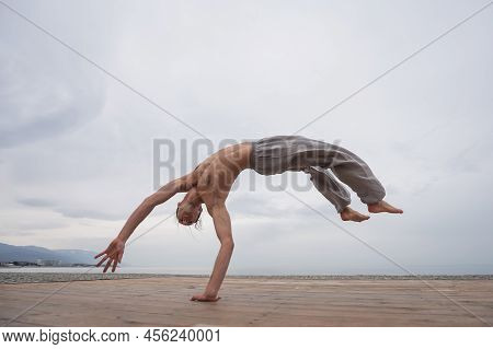 Shirtless Caucasian Man Doing Backflip On Pebble Beach.