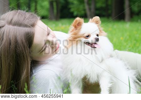Young Woman With Long Hair Lies On The Lawn In The Park With A Pomeranian Dog
