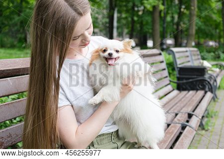 Young Woman With Long Straight Hair Sits On A Bench In Park And Holds A Fluffy Little Dog In Her Arm