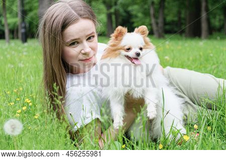 Young Woman With Long Hair Lies On The Lawn In The Park With A Pomeranian Dog