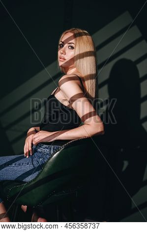 A Self-assured Woman Sits Sideways In A Leather Chair. A Painted Blonde On A Green Background With S