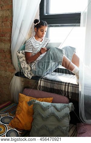 African Girl Sitting On Windowsill And Enjoying Reading On E-book During Leisure Time At Home