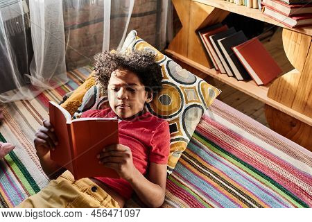 High Angle View Of African Boy Lying On Sofa And Enjoying Of Reading A Book During His Leisure Time 