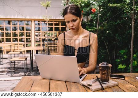 Young Female Freelancer In Trendy Outfit Sitting At Table With Cold Drink And Smartfon While Typing 