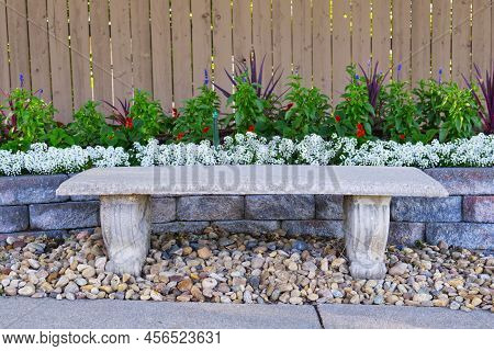 Old Vintage White Stone Bench Against Stone Wall, Wooden Fence And Blooming Flower Bed With Bright W