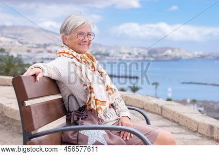 Smiling Beautiful Senior Woman With Eyeglasses Sitting Outdoors On Bench Looking At Sea Enjoying Fre
