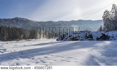 The Frozen River Is Covered With Pure Snow. Steam Rises Above The Polynya. Picturesque Boulders And 