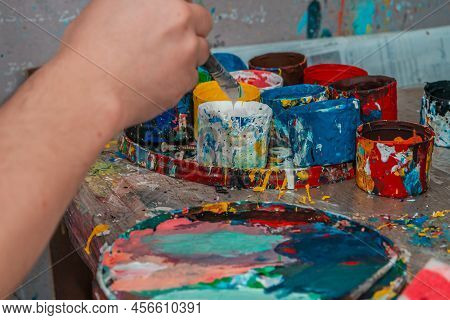 A Hand With A Brush Reaches Out To The Jars Of Paints In An Art Workshop
