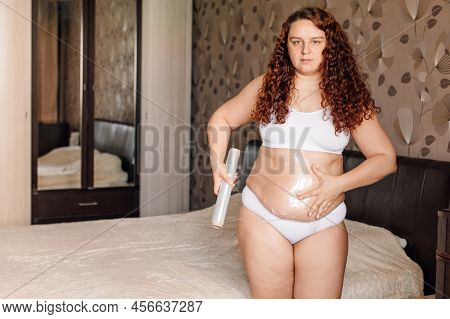 Curly Plus Size Young Woman In White Underwear Stand Near Bed In Bedroom And Wrap Sagging Belly With