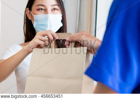 Smiling Young Asian Woman,client Accepting A Delivery Of Bag, Cardboard Box From Delivery Man Wearin